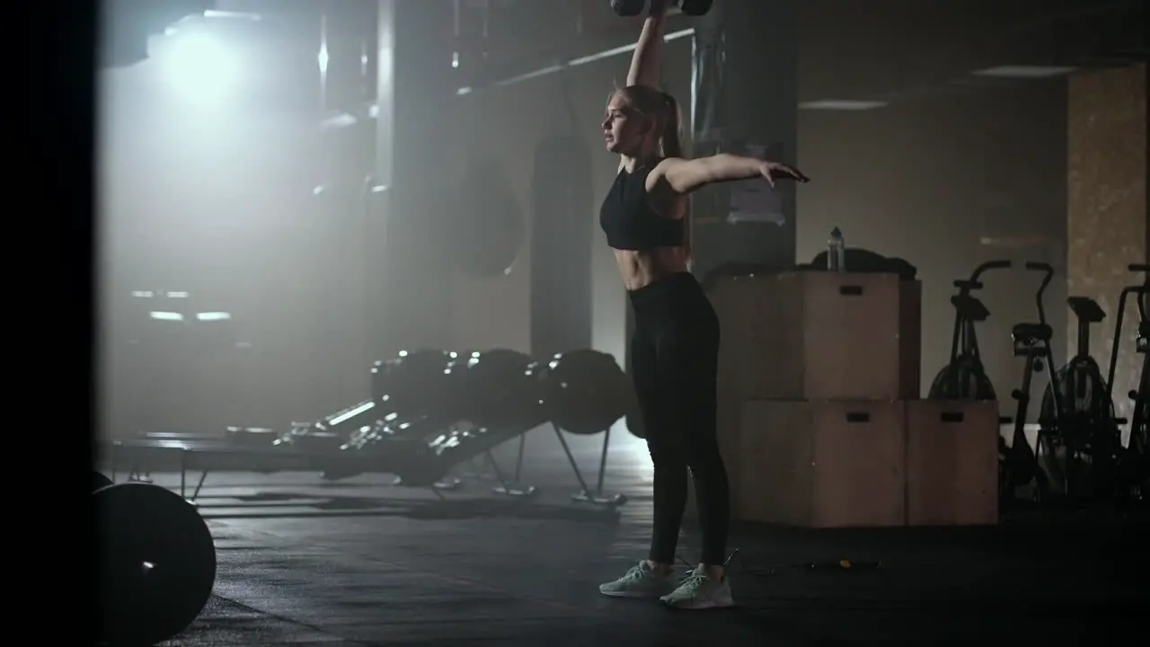 In slow motion a woman lifts a dumbbell over her head while working out in a dark gym Athletic strong woman performs a difficult workout with lifting dumbbells