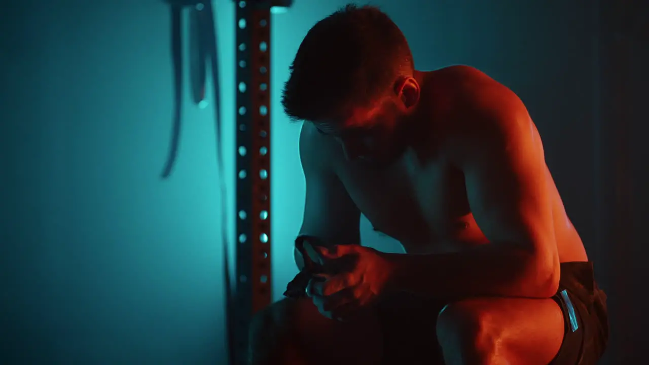 A tired man after a workout sits on a barbell and rests in the gym in blue-orange colors in slow motion