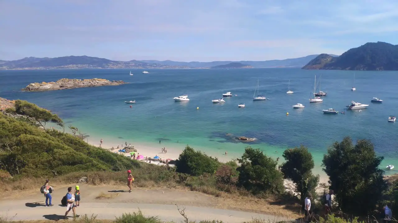 people walking in nature and the sea in the background with anchored sailboats the island in the background sunny day panoramic overhead shot turning right Cíes Islands Pontevedra Galicia Spain