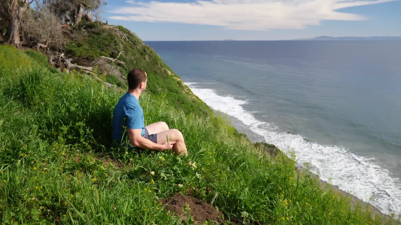 A man sitting in calming peace on the edge of a beach cliff in deep thought and meditation on a bright sunny day in Santa Barbara California SLOW MOTION