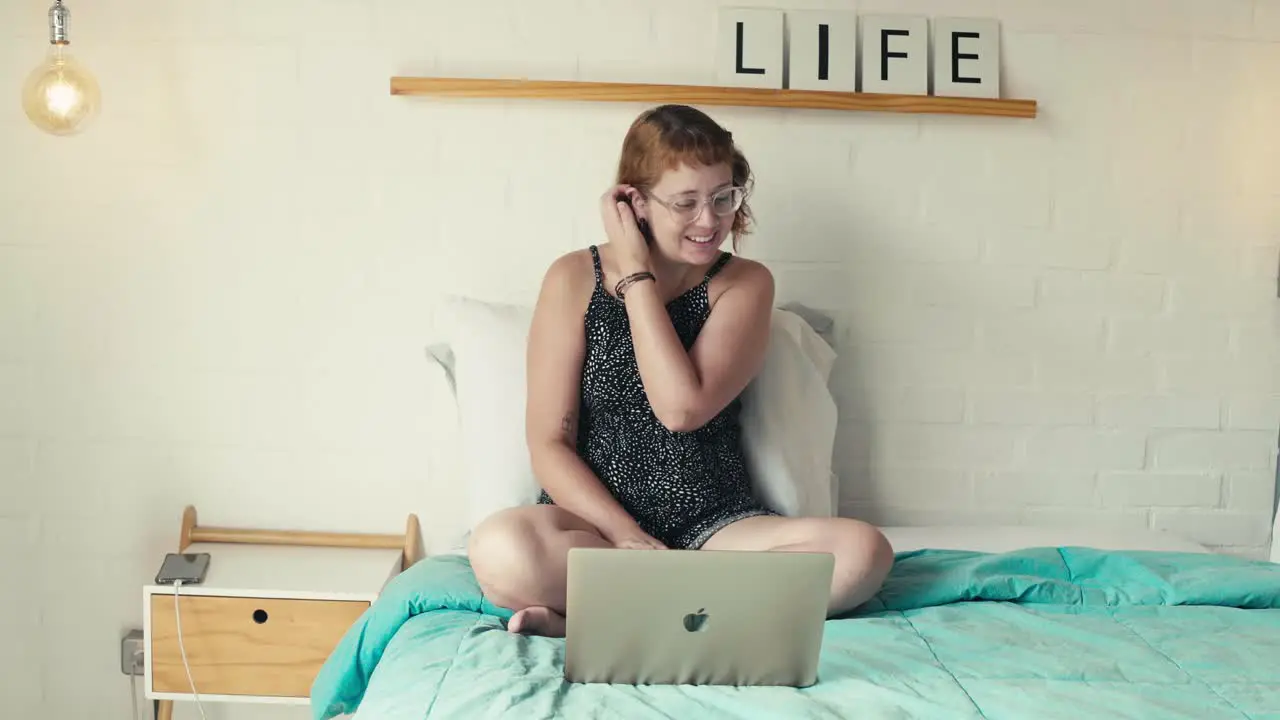 Young woman makes a video call on laptop interacts smiles sitting on bed in bright bedroom