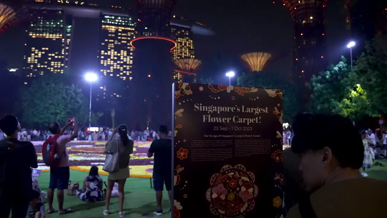 Sign Saying Singapore's Largest Flower Carpet With Visitors Taking Photos In The Background At Night Located At Gardens By The Bay