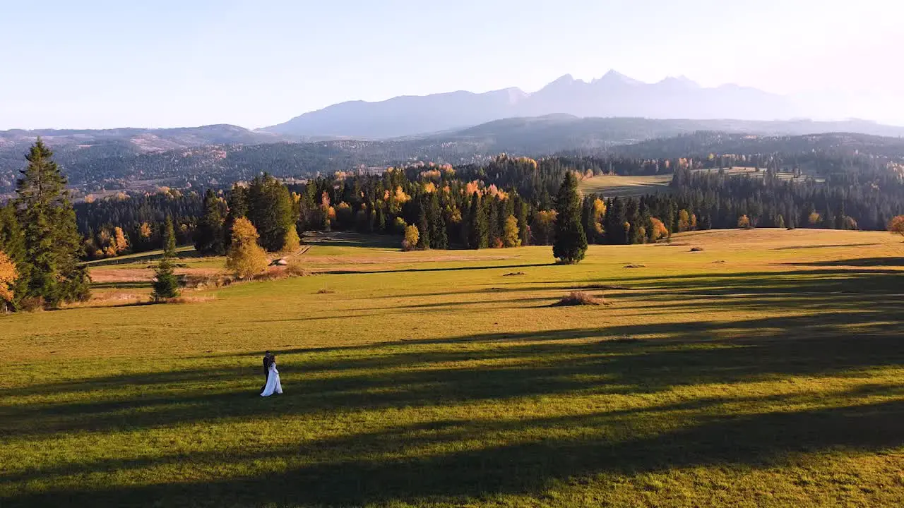 Stunning aerial shot of young couple walking through empty green field in front of huge mountains sunset forest following shot