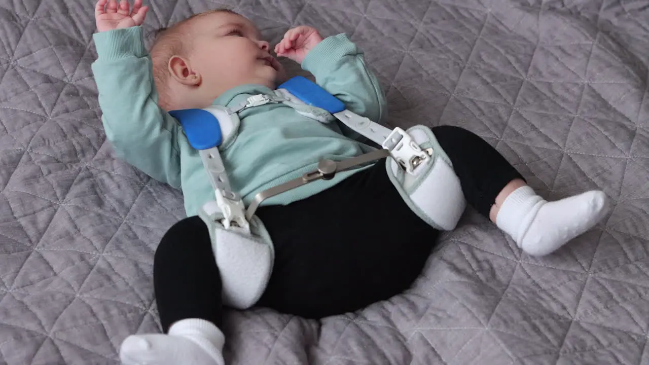 Tired baby girl wearing Pavlik Harness to correct Hip Dysplasia lies on quilt