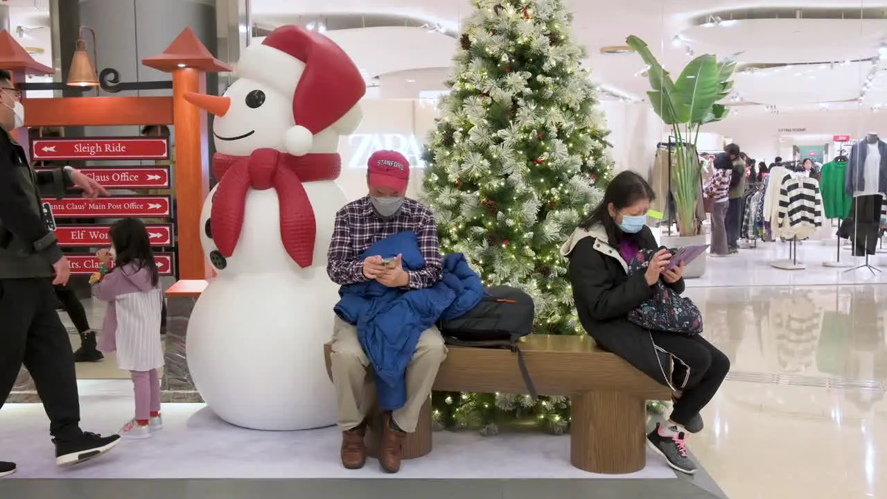 Chinese shoppers sit on a bench next to a Christmas tree and snowman installation at a shopping mall as other customers walk past them in Hong Kong