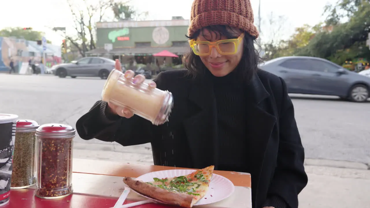 Cute Asian woman shakes parmesan topping onto margherita pizza slice at outdoor street pizza restaurant