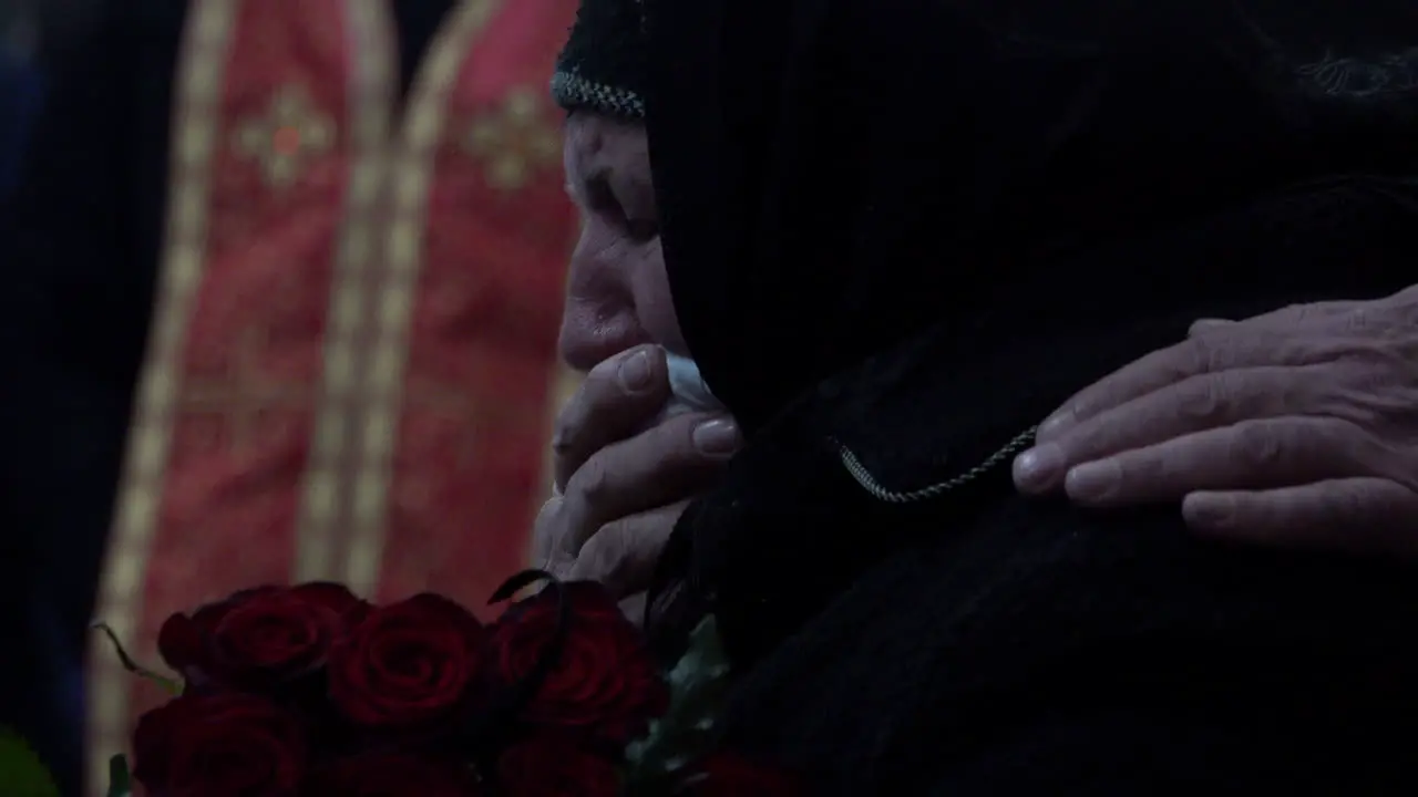 An elderly woman wearing black weeps into a handkerchief held to her mouth while holding dark red roses at a funeral of a fallen Ukraine Soldier during the Russian invasion of the country