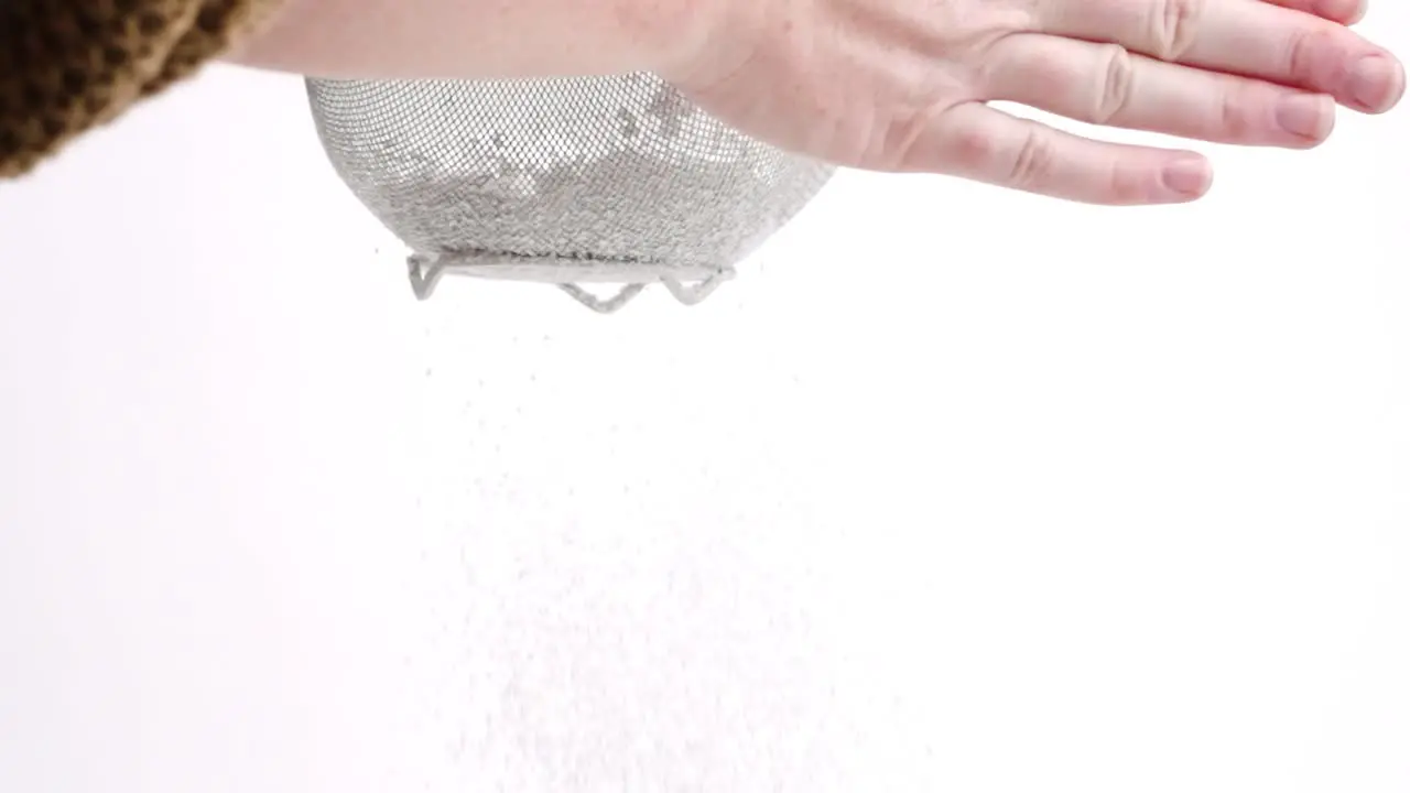 White female hand sifting powdered sugar in sieve on white backdrop in slow motion