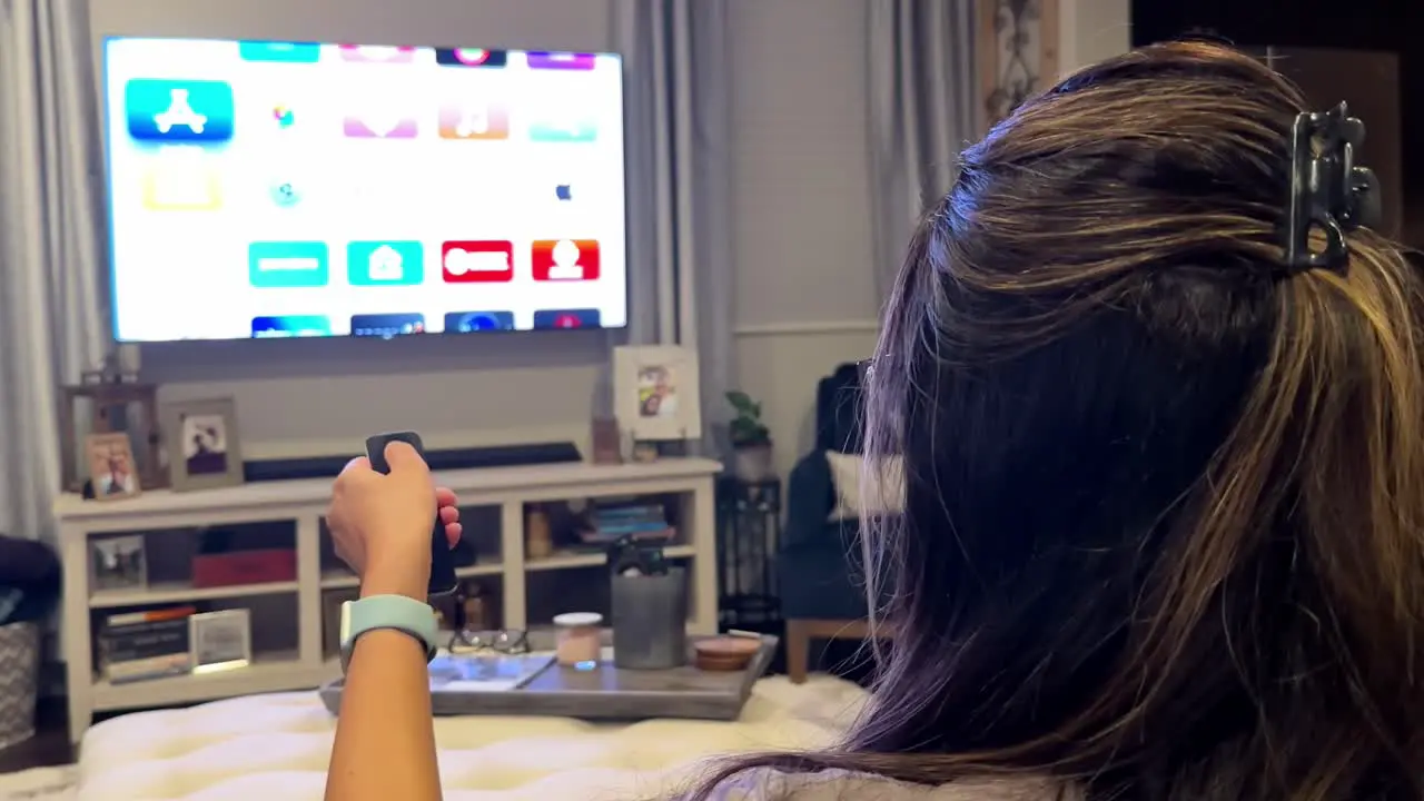 Rear view of woman scrolling streaming apps on TV using remote control at home