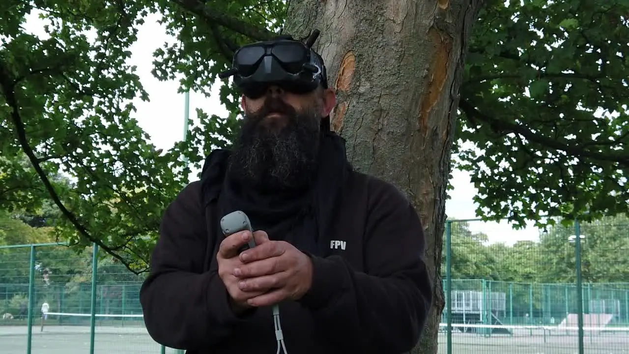 Bearded male wearing FPV virtual reality goggles controlling drone in park with motion joystick