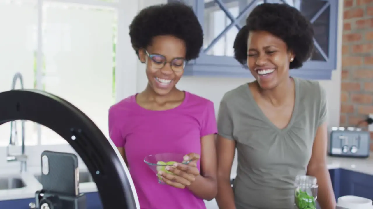 Happy african american mother and daughter preparing healthy drink making video using smartphone