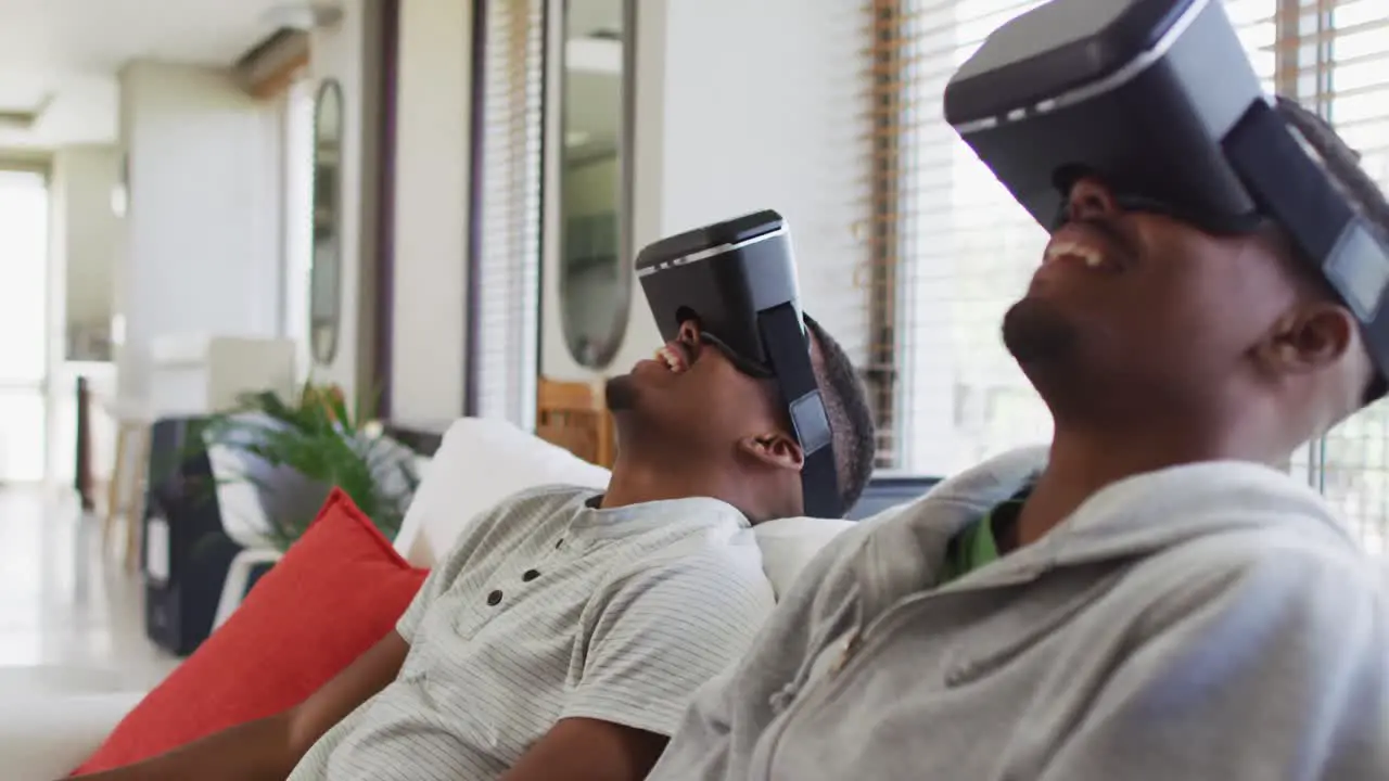 Two african american teenage twin brothers sitting on couch using vr headsets and smiling