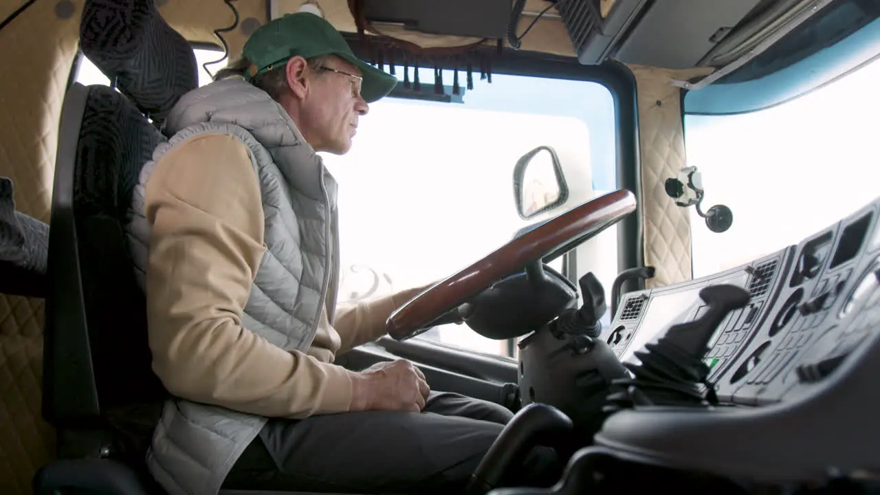 Side View Of Older Worker Wearing Cap And Vest Driving A Truck In A Logistics Park 5