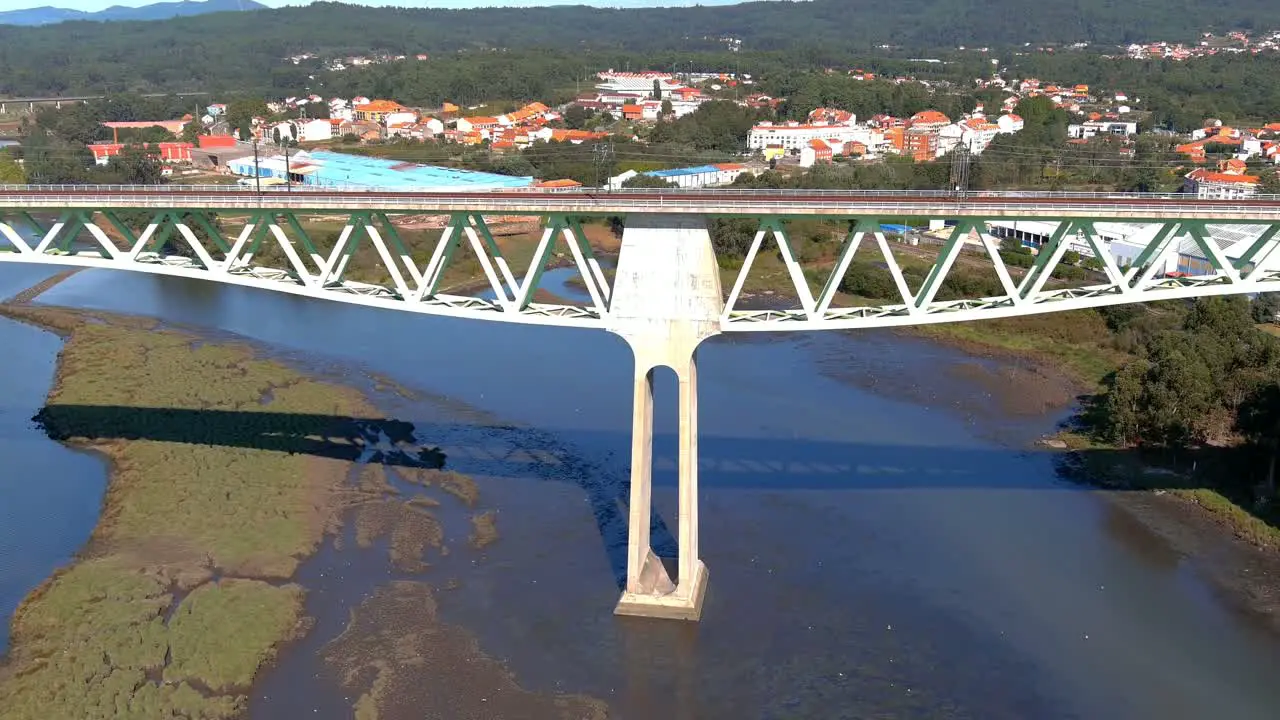 Railway bridge with power lines over the ulla river with low water level seagulls flying low industrial buildings behind shot of rolling overhead drone traveling forward Catoira Galicia Spain