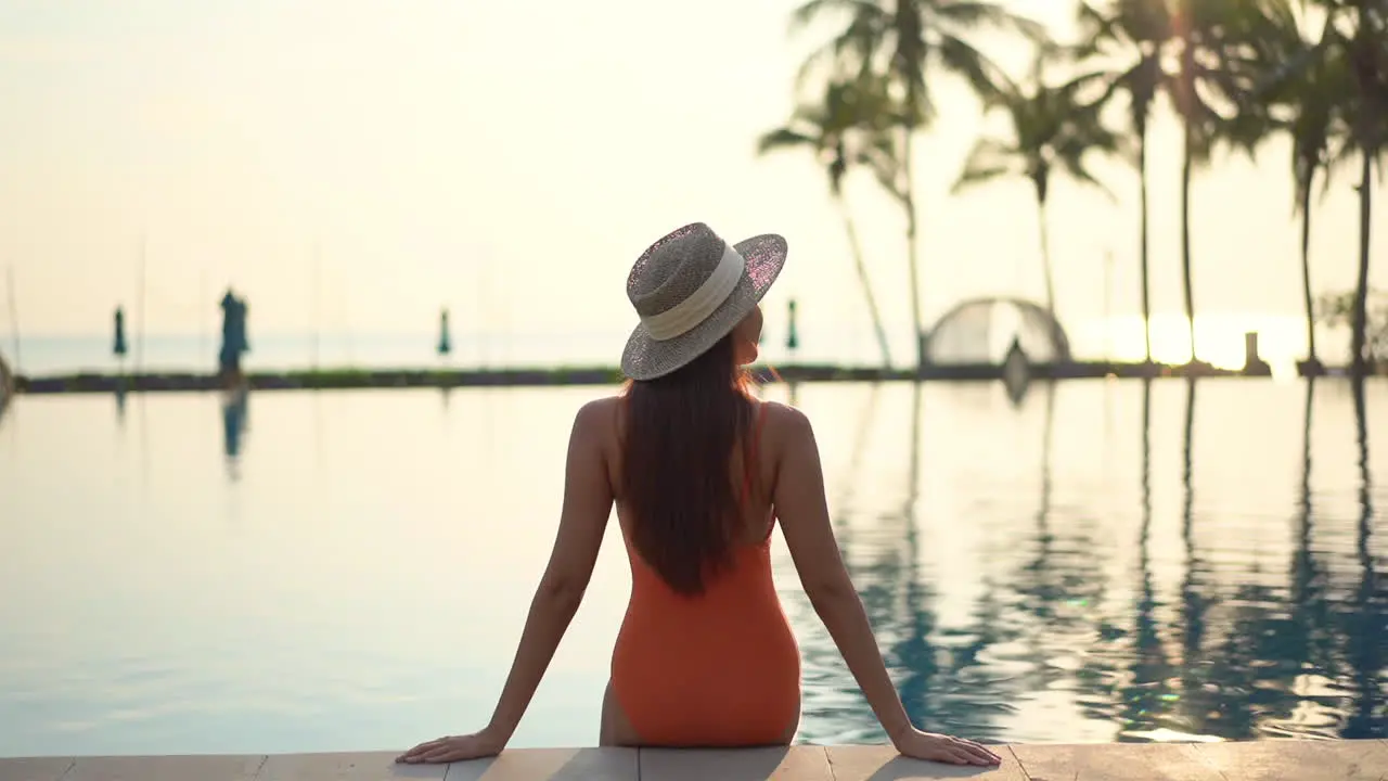 Back view of Fit female model wearing an orange monokini swimwear suit and hat sits on the edge of the swimming pool leaning on arms at sunset