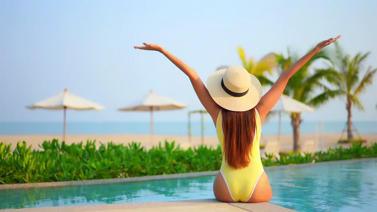 Back of petite female in yellow swimsuit raising hands sitting by the pool with beautiful view of beach and parasols of luxury beachfront hotel resort full frame