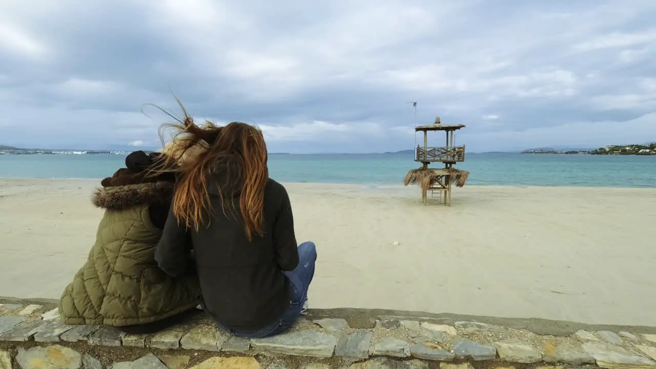 Two women in winter are sitting on the wall at the beach