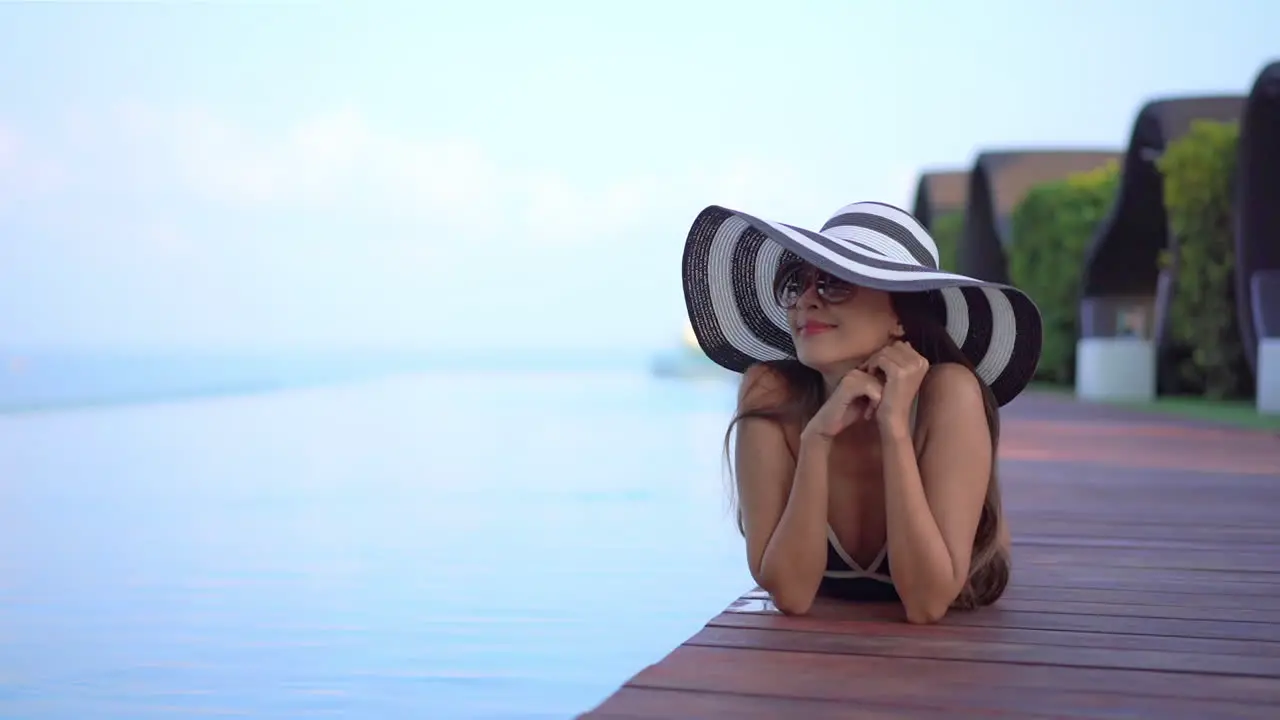 Asian smiling woman with large striped hat and vintage sunglasses sunbathing lying poolside