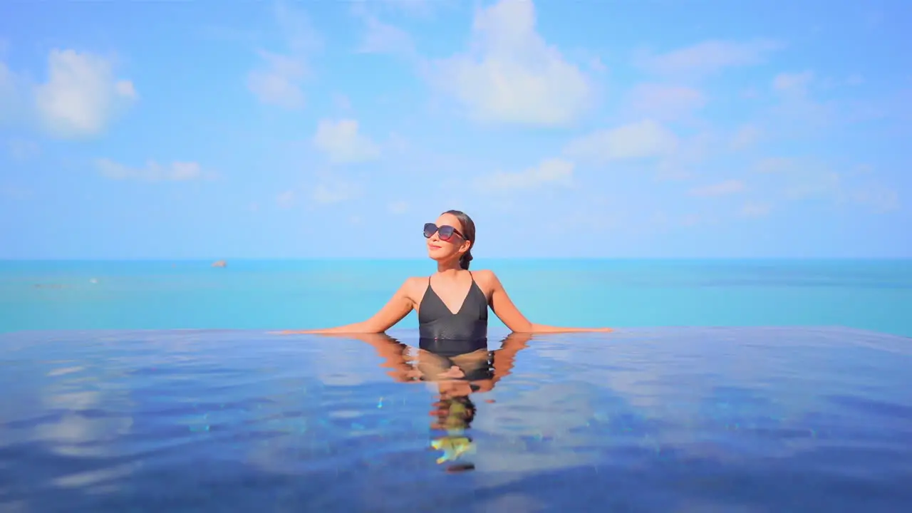 An attractive young woman leans along the edge of an infinity-edge pool with an ocean horizon in the background