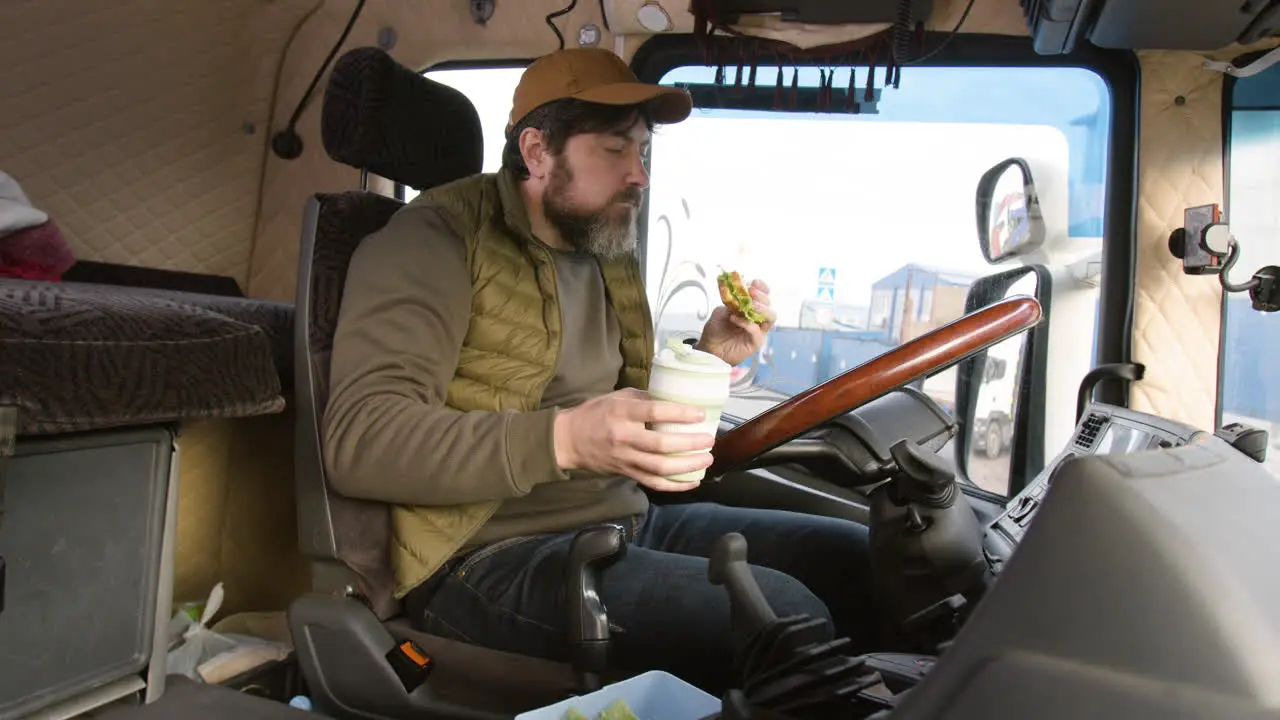 Side View Of Worker Sitting In A Truck In A Logistics Park While Holding A Sandwitch And Drinking