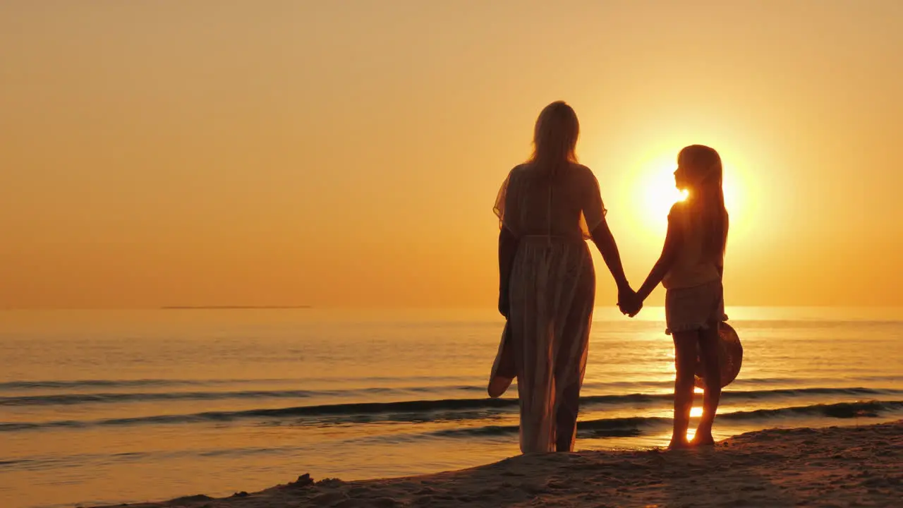 A Woman Holds A Child's Hand Stand Together On The Seashore Look At A Beautiful Sunset Mother With D