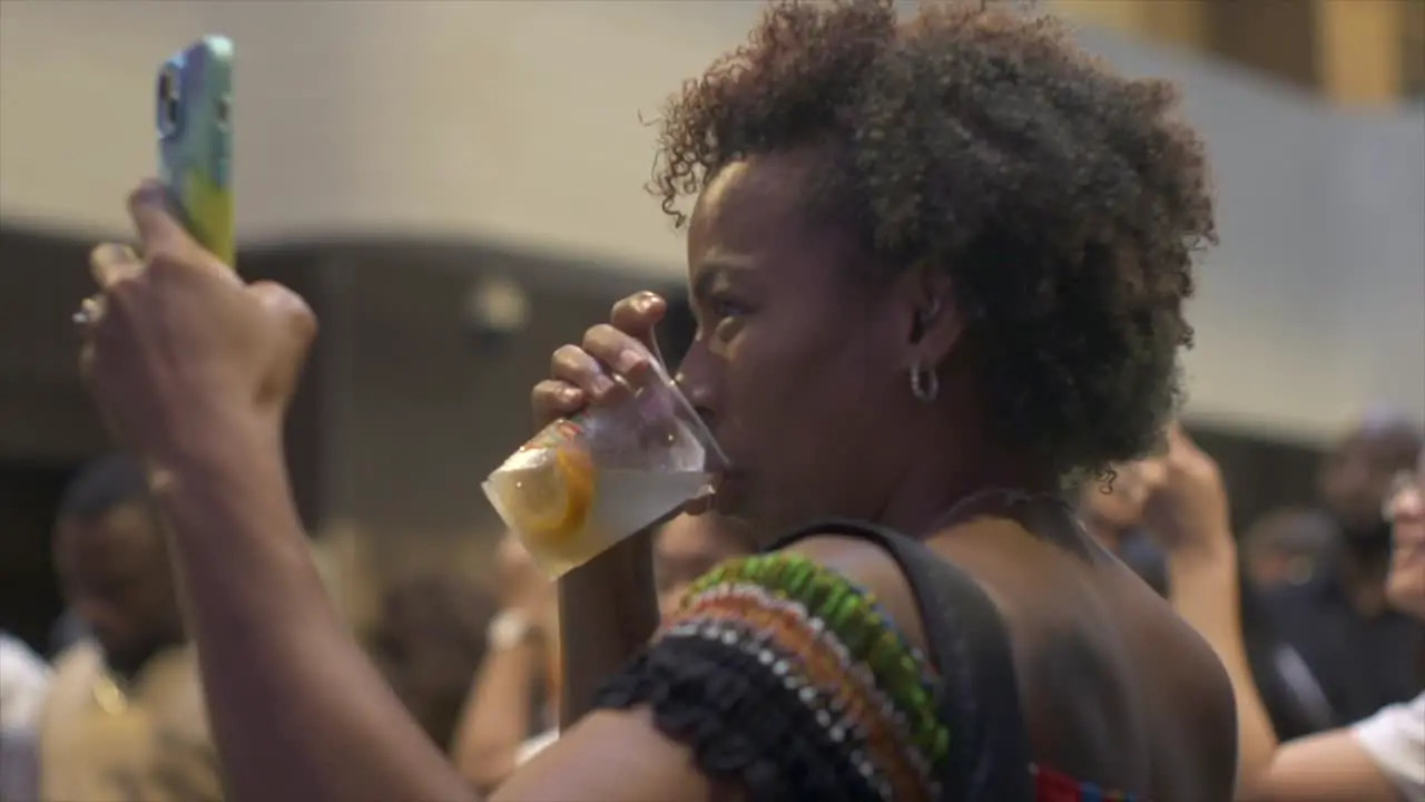 African woman drinking alcoholic beverage while using cellphone to film and live stream at outdoor ethnic cultural event filmed as close up in slow motion