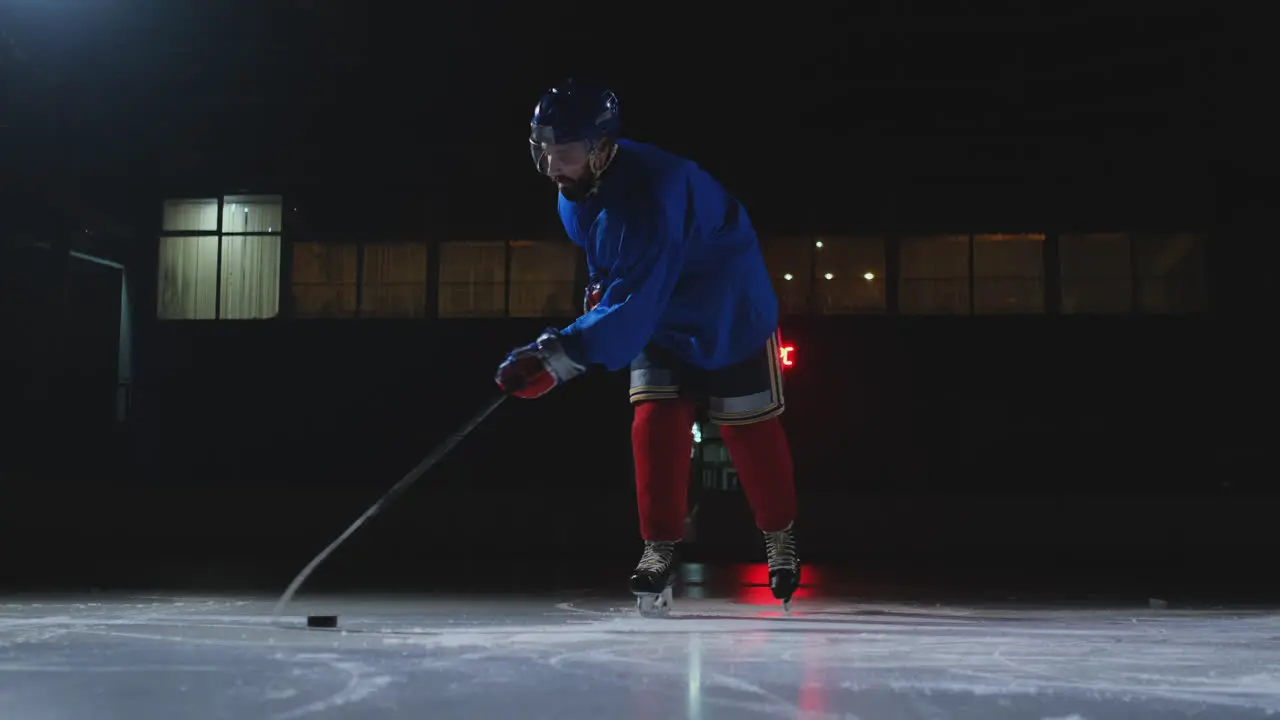 Man hockey player with a puck on the ice in hockey form leaves with a stick in his hands out of the darkness and looks straight into the camera