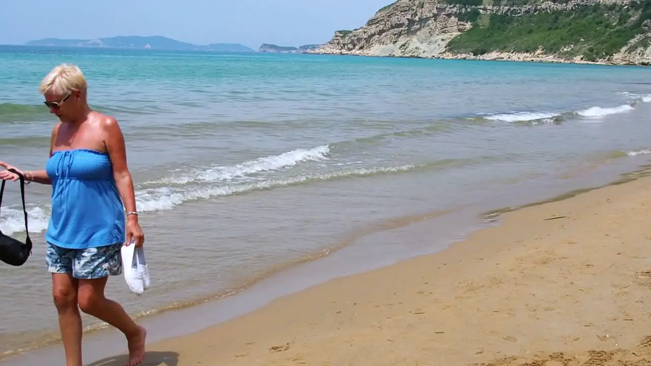 Attractive middle aged female walking barefoot on beach feeling hot during midday sun Corfu Greece