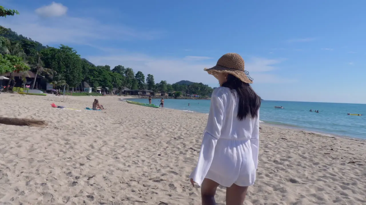 Woman in a white dress and brown hat walks on a beautiful sandy beach