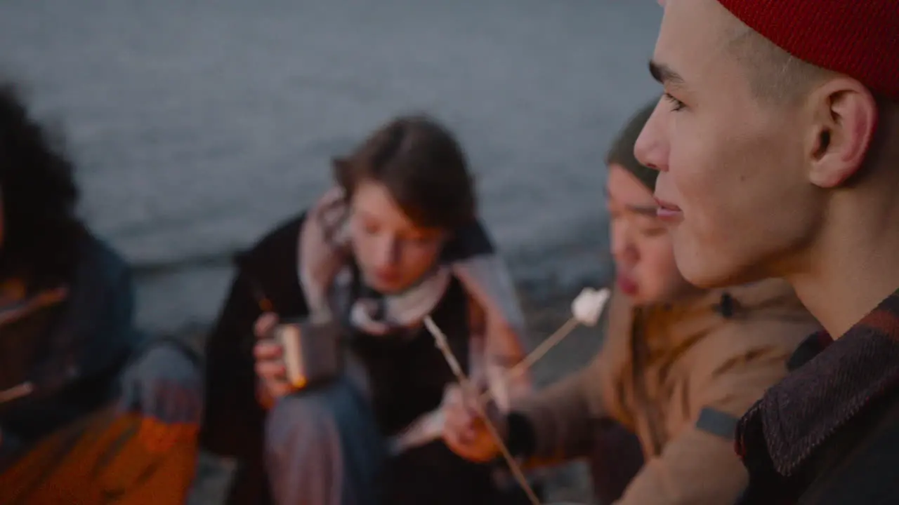 Rear View Of A Teenage Boy Roasting And Eating Marshmallows Sitting Around Bonfire With Her Friends