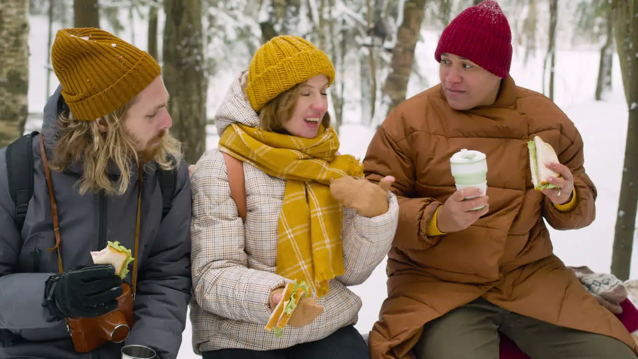 Three Friends Sitting On A Tree Trunk Talking And Eating In A Snowy Forest