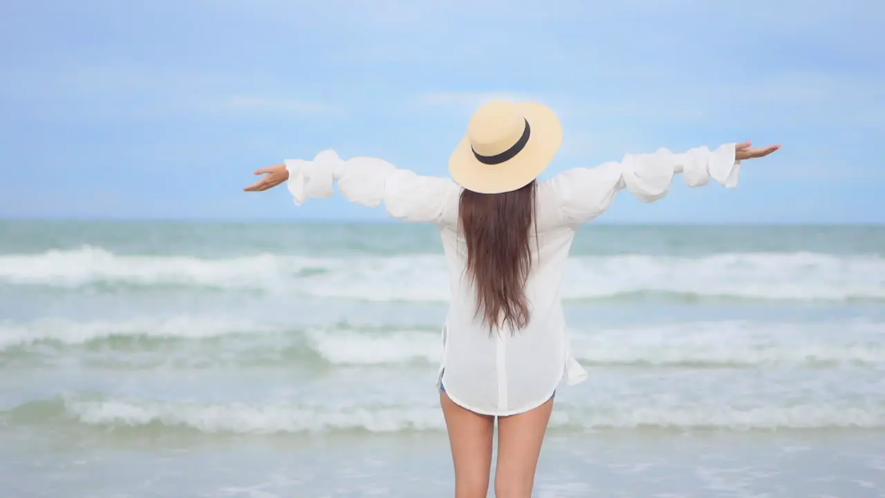 Joyful woman with long hair and floppy hat raising and spreading hands in front of waves and horizon of tropical sea back view full frame slow motion