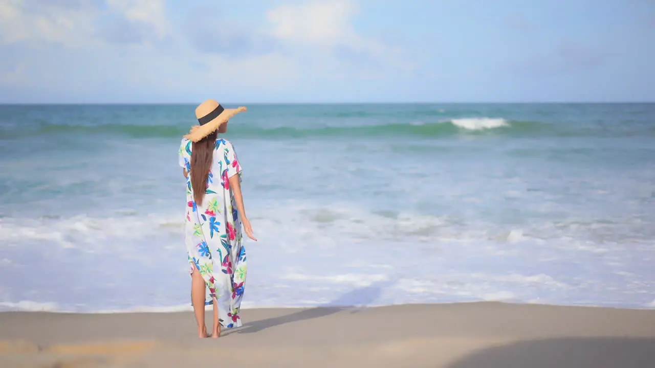 Lonely Stylish Female in Colorful Summer Dress and Hat Standing on Sand in Front of Tropical Sea Waves Back View