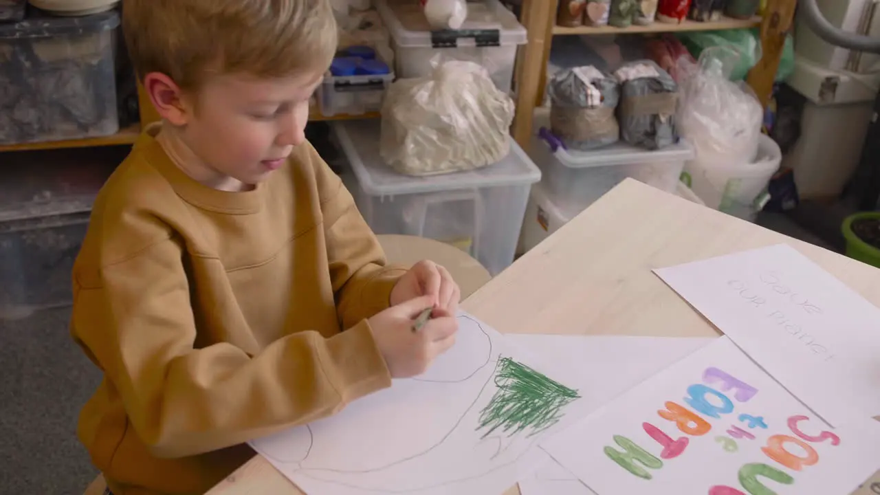 Blond Kid Drawing With Green Pencil Sitting At A Table In A Craft Workshop Where Is Signs With Environmental Quotes