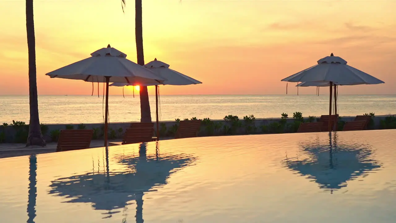 Orange tropical sunset above calm sea horizon swimming pool and beach umbrellas with mirror reflection full frame