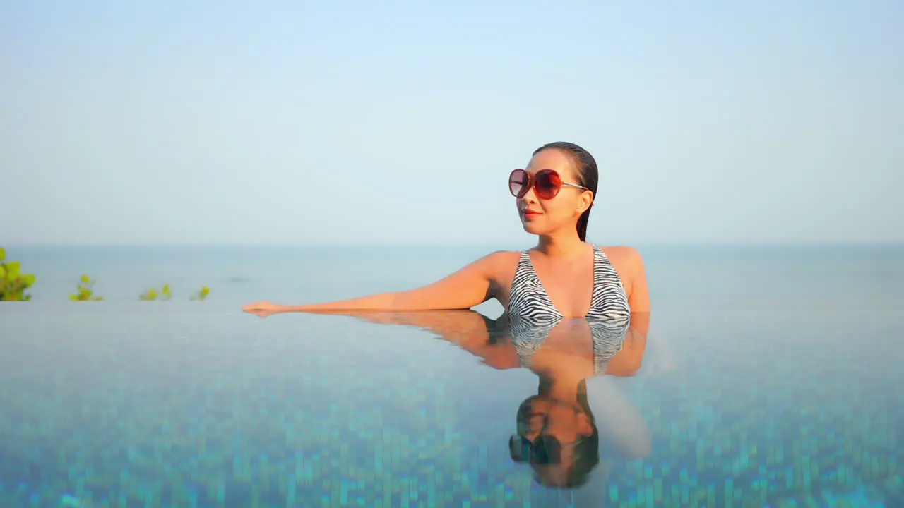 Sexy happy asian woman in infinity swimming pool with sea horizon in background mirror reflection on water on sunny day at exotic tropical location