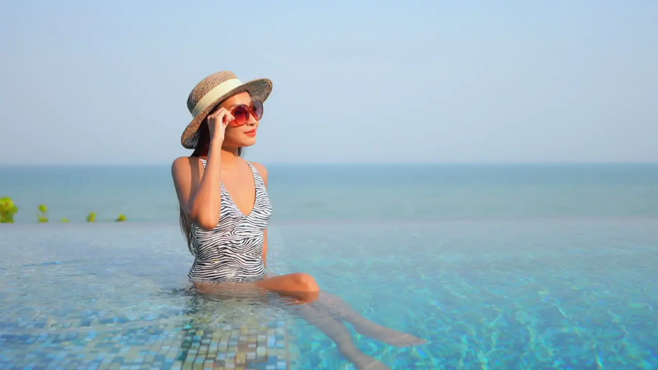 Charming Asian woman sitting in infinity pool water contemplating panorama