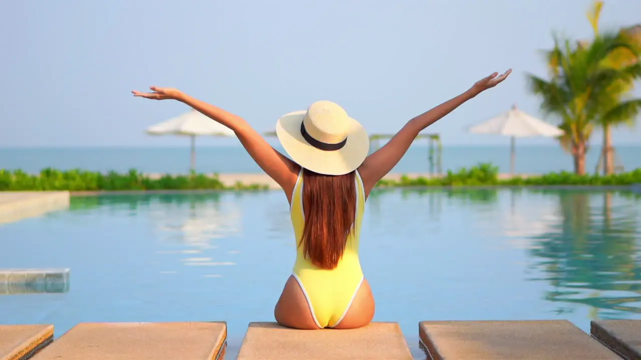 Back view of woman with large hat and yellow swimsuit sitting on pool edge and raising arms to sky