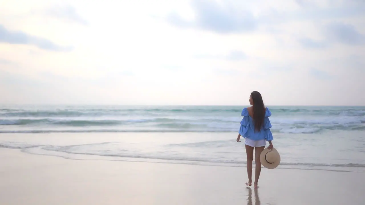 Back to the camera slow motion rule of thirds an attractive woman in shorts and a bohemian blouse holds a sunhat in one hand as she walks into the oncoming surf