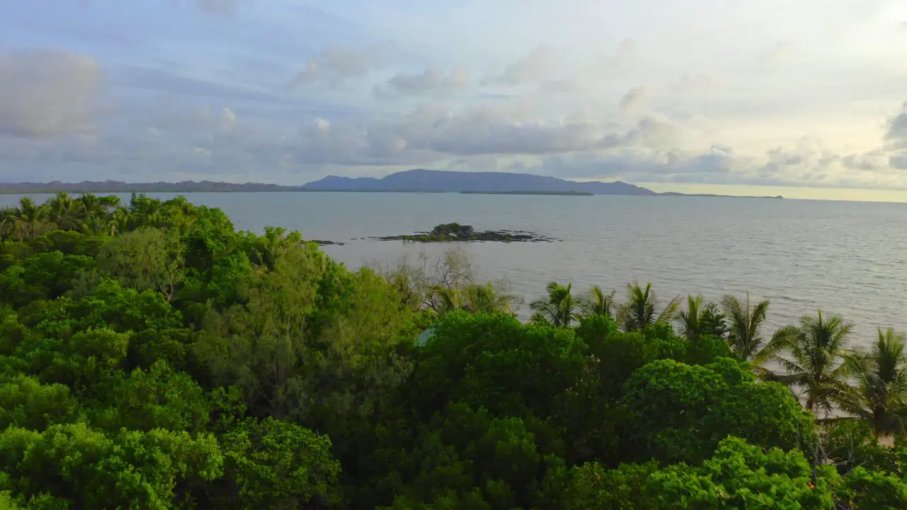 fly over a little tropical wood toward a little rock formation in the sea near a beach where some people are hanging out during a warm day