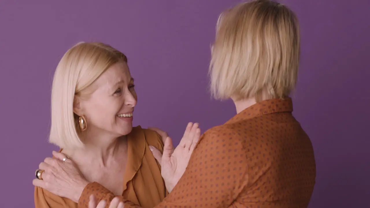 Dos Mujeres Rubias Maduras Con Pantalones Marrón Abrazándose Y Sonriendo Con Fondo Morado