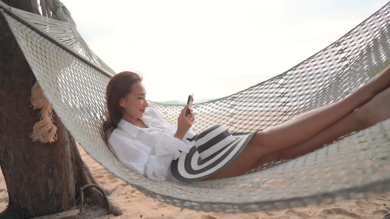 Young asian woman laying on beach hamcock and looking at smartphone with smile