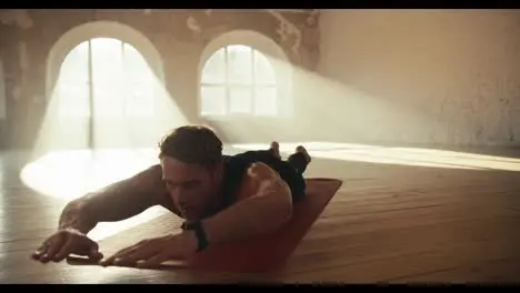 A male athlete in a black sportswear summer uniform does low boat exercises in a brick sunny gym