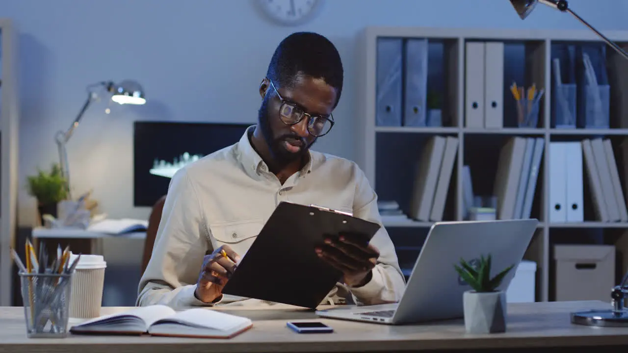 Office Worker Being Still Sitting At Desk Working With Some Documents With A Pencil In The Office At Night