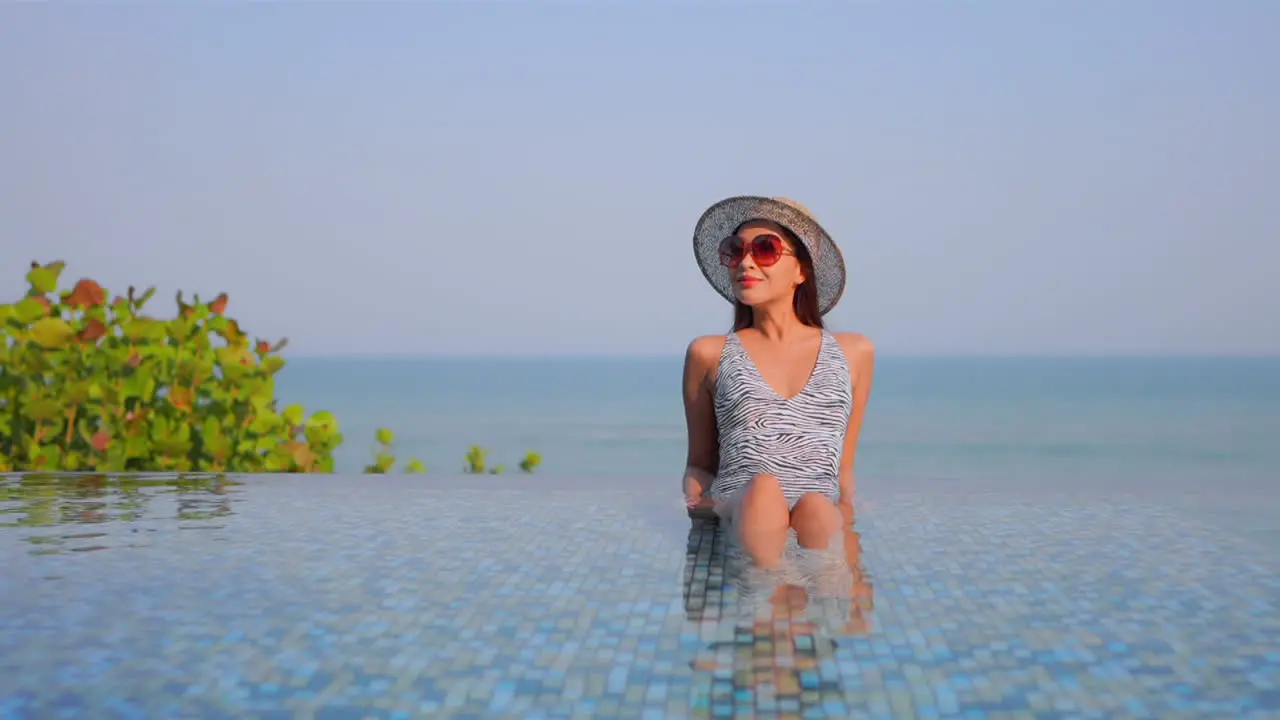 Stunning girl sitting leaning on elbows in shallow water inside infinity pool enjoying gorgeous tropical scenery daytime slow-motion