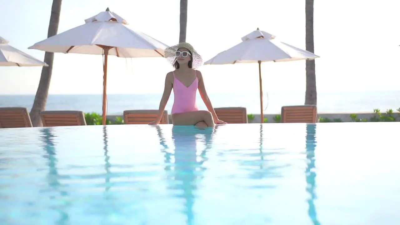 Fashionable Asian woman sitting on infinity swimming pool border enjoying in warm tropical weather