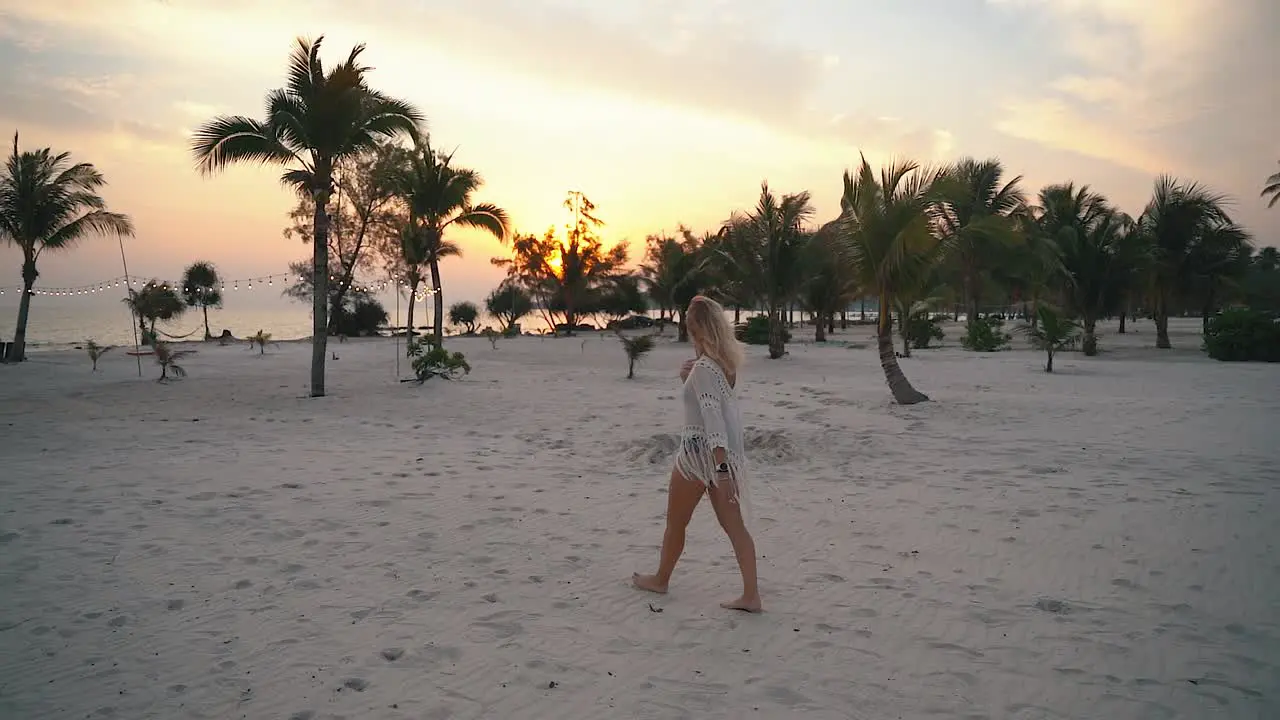 Young blond woman in summer clothes walks barefoot on the white beach among the green palm trees during a beautiful sunset