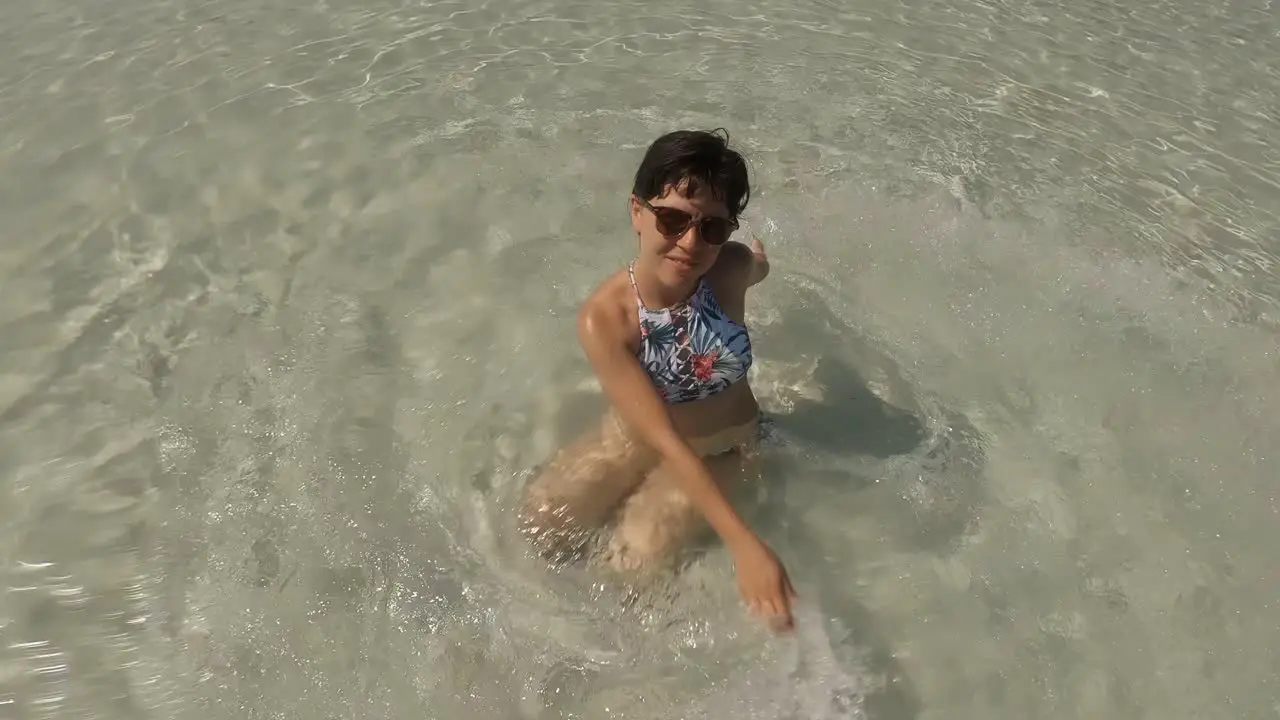 A happy girl in swimming suit playing around with her hands in the sea while sitting at the bottom of the white sand