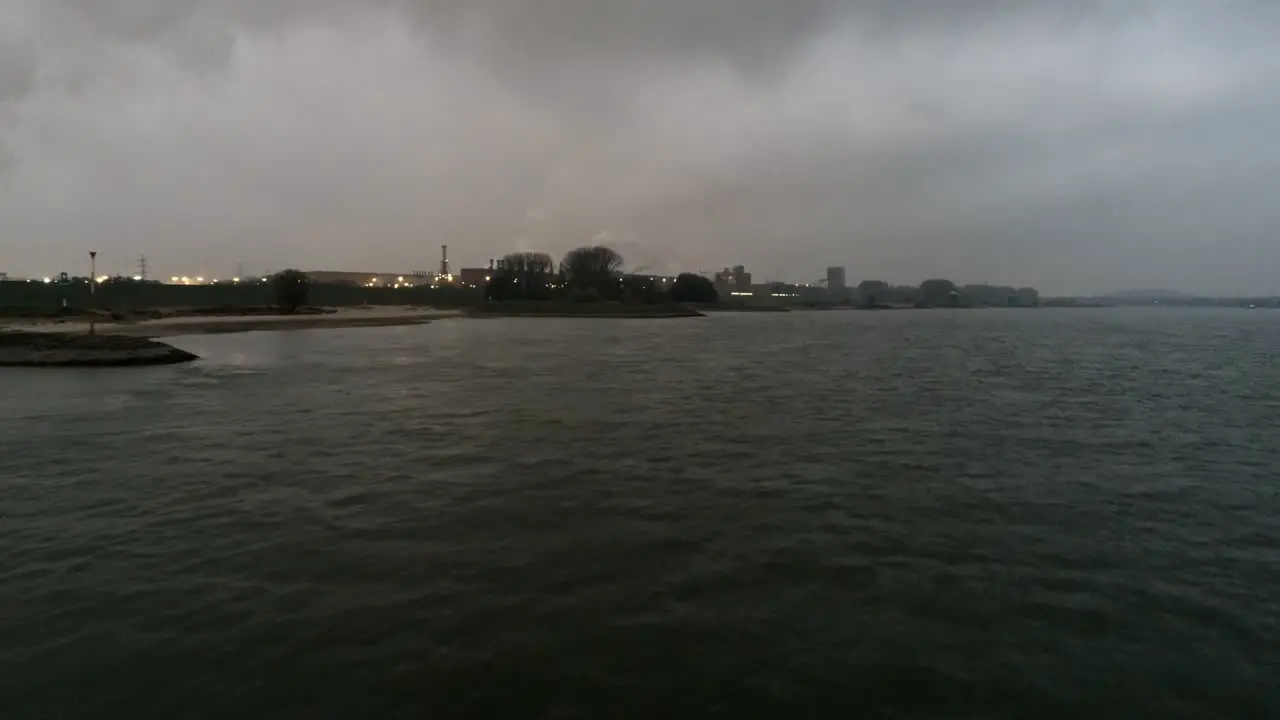 Drone flying above the water surface towards the industrial zone during nightfall on an overcast day