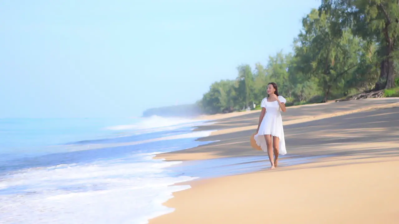 A young fit woman in a white sundress walks along the beach while the waves roll in